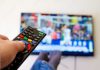 FCC to crack down ‘junk fees’ on cable and satellite TV, requiring to display ‘all-in’ price Close-up macro of man's hand with TV remote control watching a soccer match