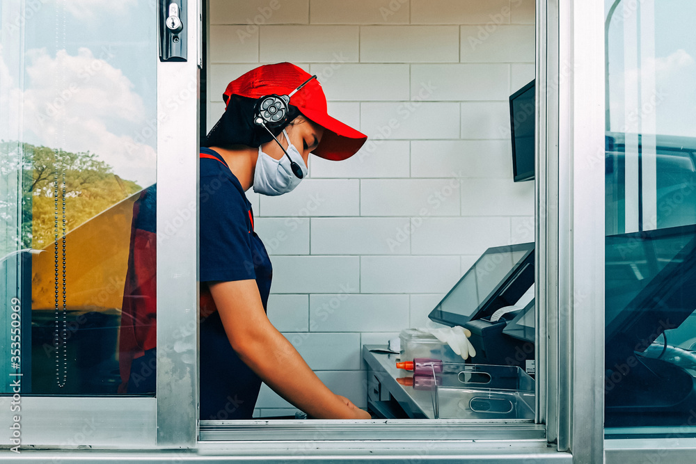 Bangkok, Thailand - May 28, 2020 : fast food cashier in drive thru service wearing hygiene face mask to protect coronavirus pandemic or covid-19 virus outbreak working on counter at the station.