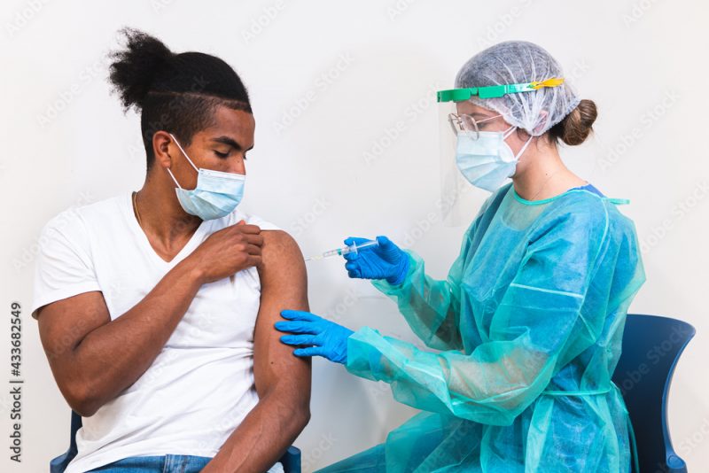 Female medical specialist in protective uniform, latex gloves and face mask vaccinating African American man patient in clinic during coronavirus outbreak