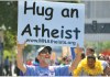 They Preach Against God Online — Some ‘Zealous Atheists’ Still Pray Man holding a cardboard sign reading “Hug an Atheist,” symbolizing empathy and connection within the Zealous Atheists study on nonreligious communities.